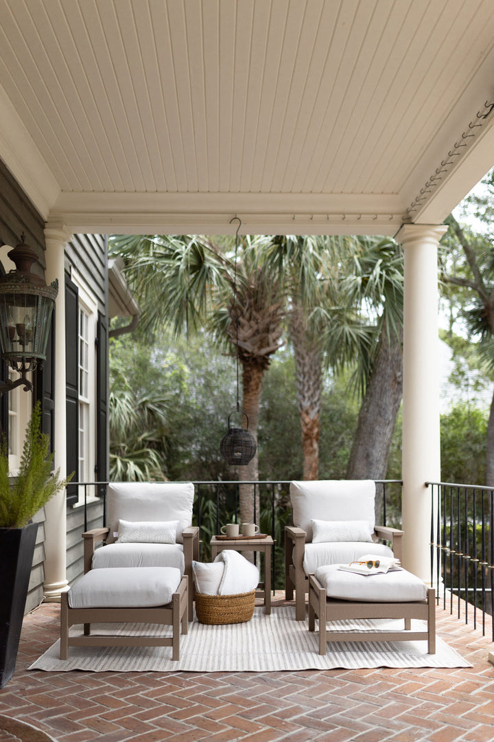 Cozy patio furniture arrangement under a covered porch with two wooden club chairs, ottomans, and a side table, accented with white cushions and a woven basket on a brick floor, surrounded by lush greenery.