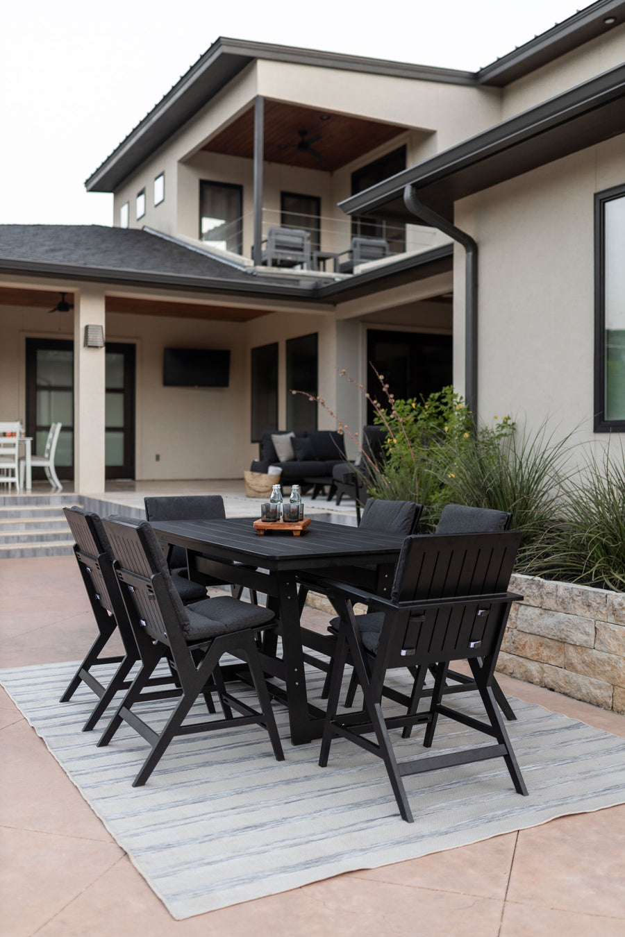 Modern black outdoor dining furniture set on a stone patio with a striped area rug, featuring a sleek table and six chairs, in front of a contemporary two-story house.