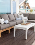 classic-Sectional in Natural Teak with cushions in Play Mushroom, complemented by throw pillows in various shades including Canvas Skyline and Palm Springs Sand, paired with a white coffee table on a wooden deck overlooking a coastal view.